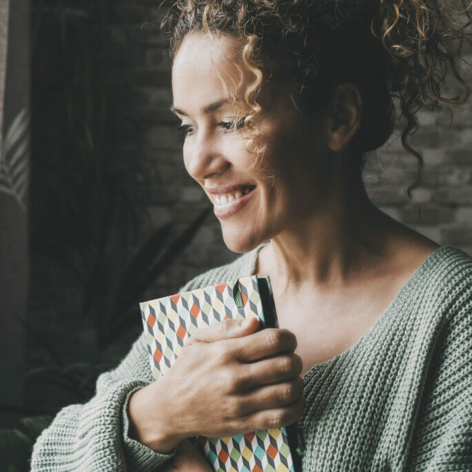 Happy smiling expression woman hold book and look outside the wi Happy smiling expression woman hold book and look outside the window at home. Dark background. Joyful face portrait of female people in indoor leisure activity alone. Attractive middle age lady indoor